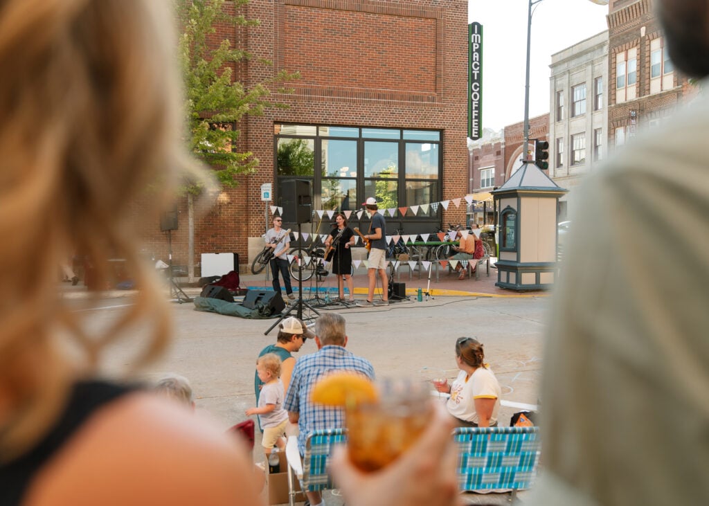 A camera peers over a couple's shoulder to view musicians playing at Lawn Chair Night in Downtown Decorah.